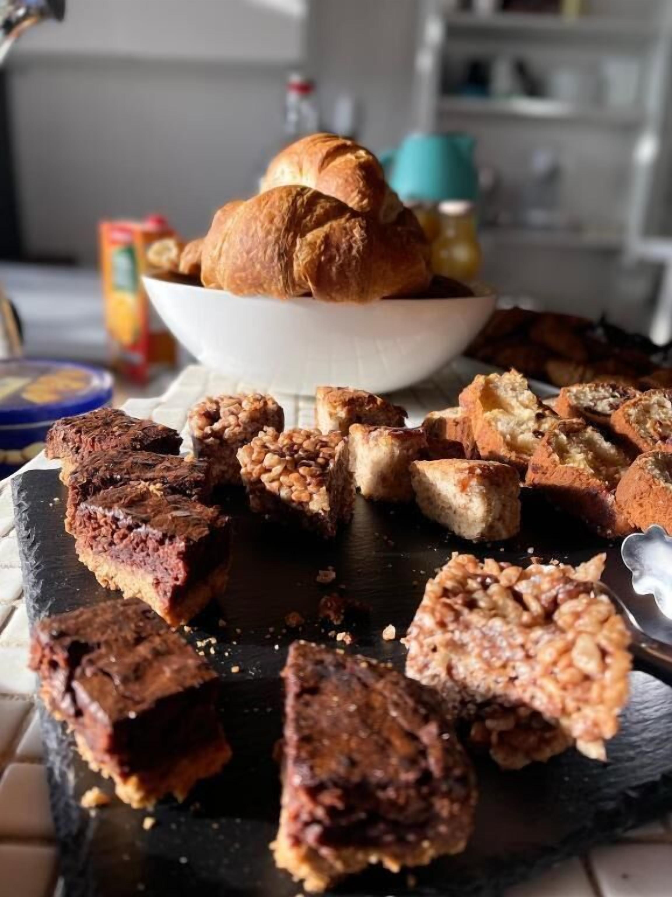 Variedad de productos horneados sobre una tabla de pizarra, incluyendo un bol de cruasanes y varios tipos de pan y pastelería.