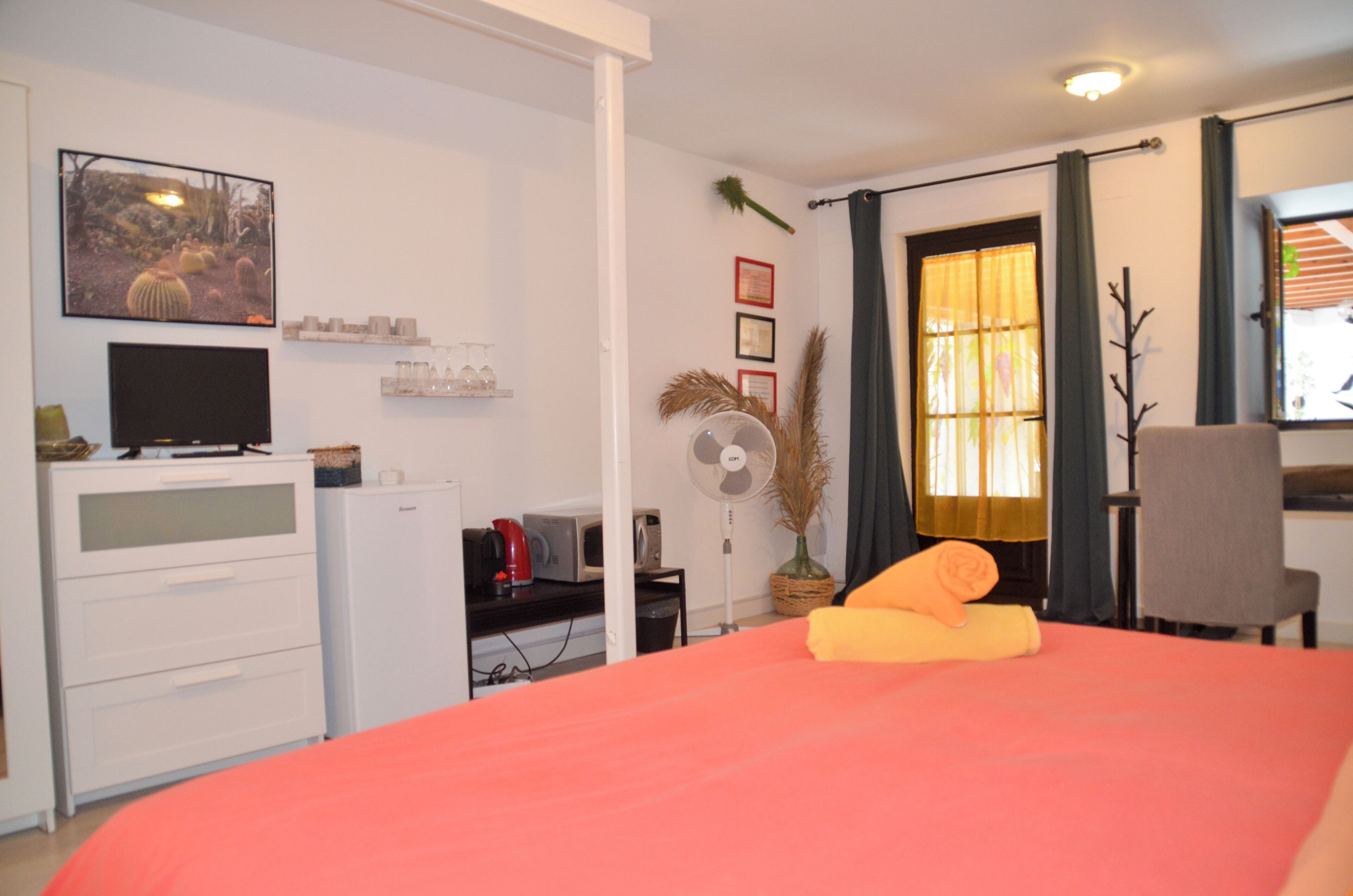 Quarto de hotel com cama, escrivaninha, cadeira, televisão, ventilador e janela com cortinas.