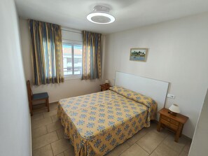 Ein Schlafzimmer mit Bett, Stuhl, Nachttisch, einem Fenster mit Vorhängen, einer Deckenleuchte und einem Wandbild.