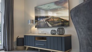 A modern living room with a large flat-screen TV mounted on the wall, a dark grey entertainment unit, a lamp, and a decorative sign. - House Munkmarscher Straße 14c (Westerland)