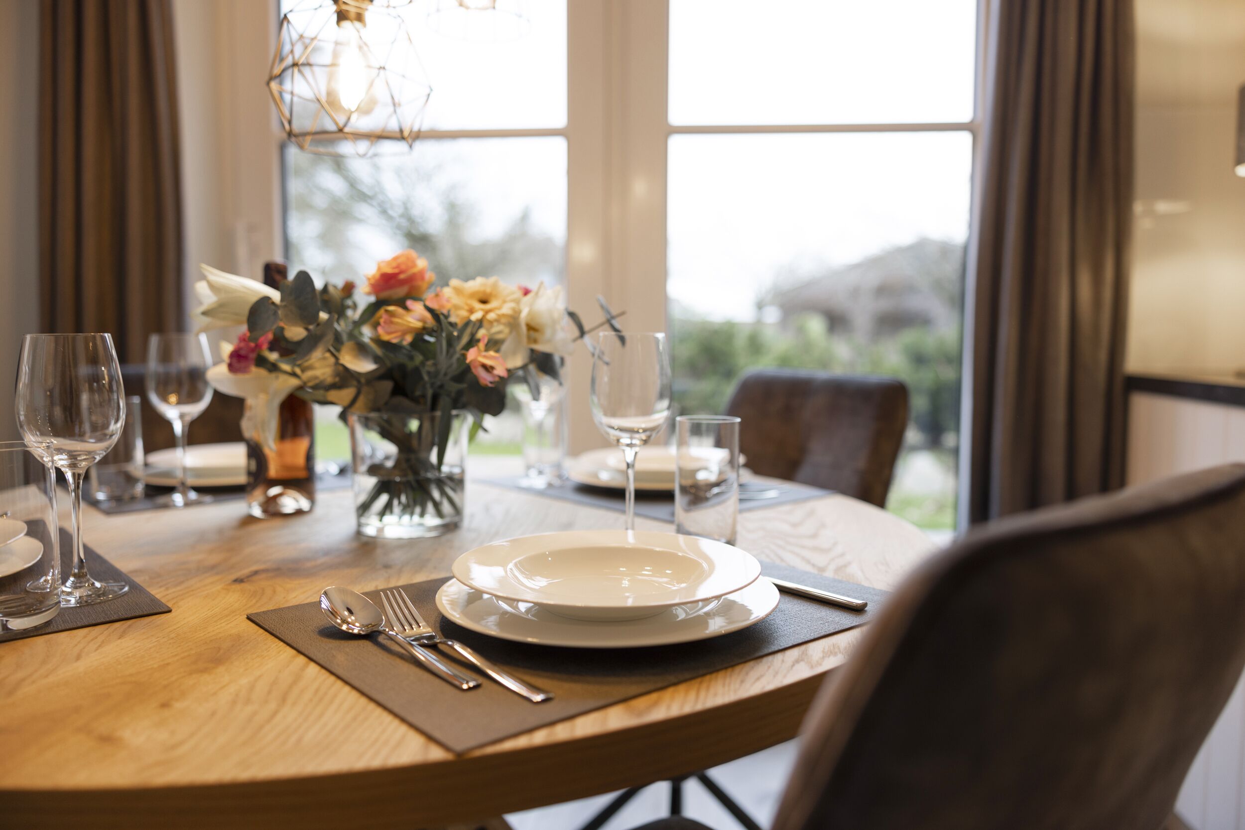 A dining table set with plates, cutlery, and glasses, with a vase of flowers and a view of greenery outside.
