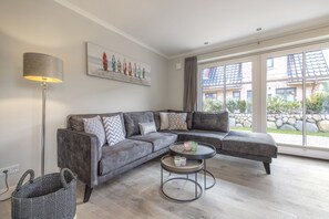 A modern living room with a grey sectional sofa, a round coffee table, a floor lamp, and a large window with a view of a building and greenery.