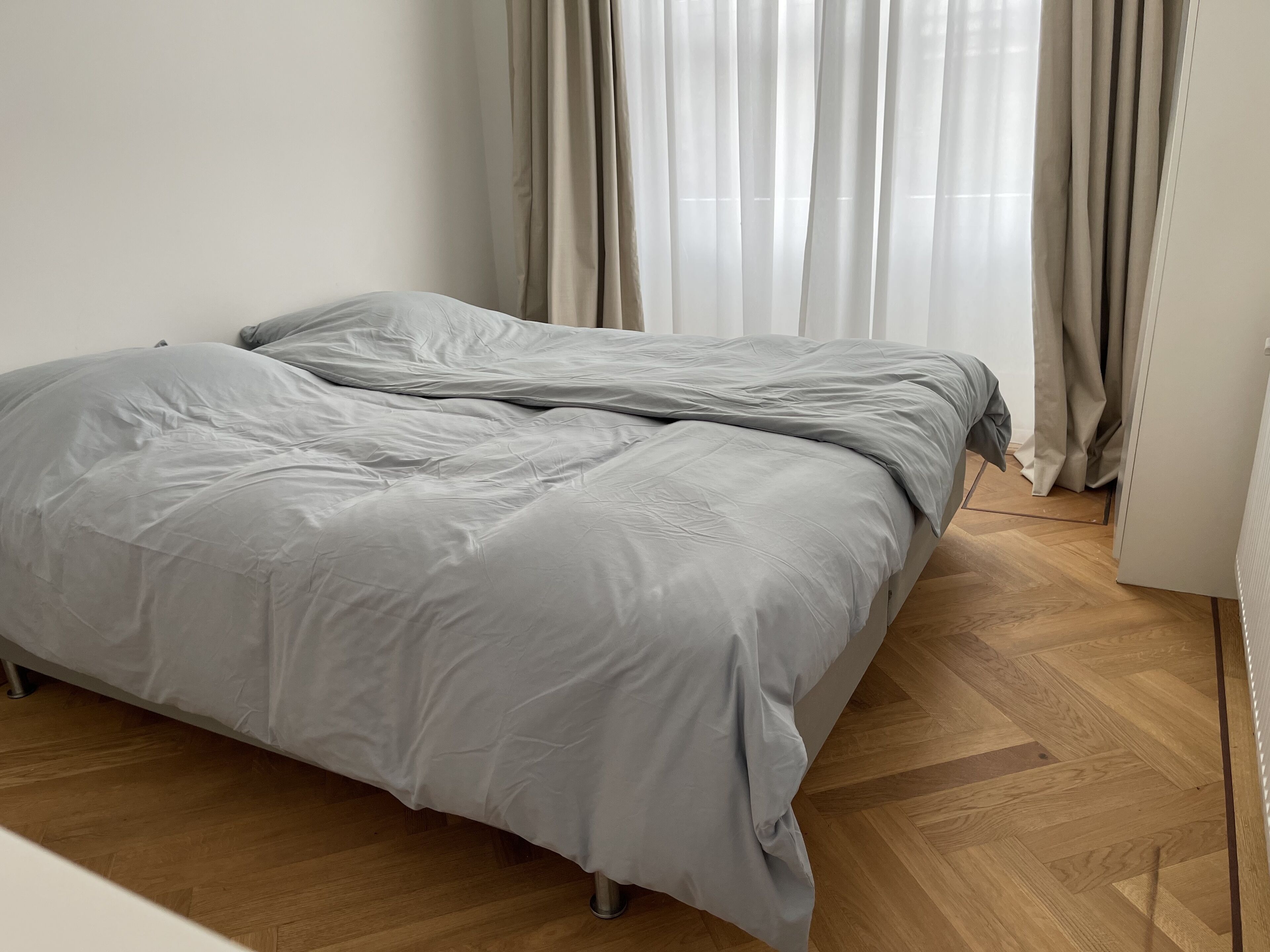A bed with a grey duvet in a room with wooden flooring and beige curtains.