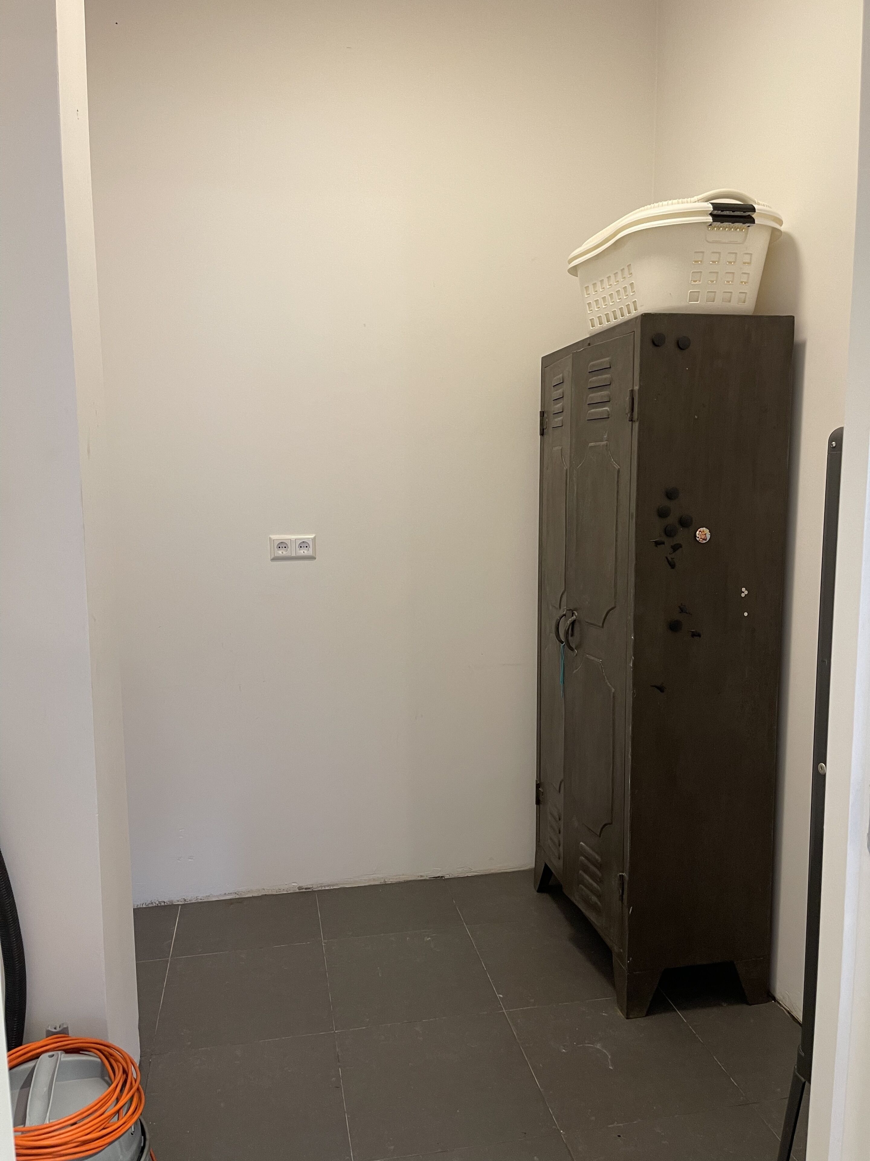 A small, dimly lit room with a wooden cabinet, a power outlet, and a coiled cable on the floor.