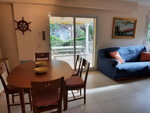 A dining area with a round table and chairs, a blue sofa with cushions, a painting on the wall, and a view through a glass door to an outdoor balcony.