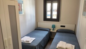 Une petite chambre propre, avec deux lits simples, une fenêtre, une porte et une petite table avec une lampe.