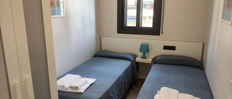 A small, clean room with two single beds, a window, a door, and a small table with a lamp.