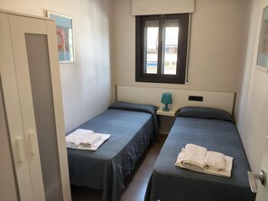 A small, clean room with two single beds, a window, a door, and a small table with a lamp.