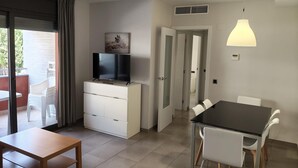 Une chambre moderne avec un téléviseur posé sur un meuble blanc, une table à manger avec des chaises et une grande fenêtre avec des rideaux.