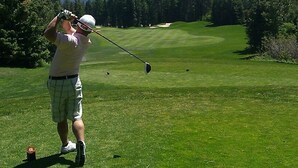 Un terrain de golf avec un joueur en plein élan, un mât de drapeau et un ciel dégagé.