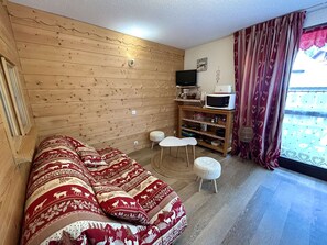 A cozy room with a wooden wall, a sofa, a TV, a small table, and a window with curtains.