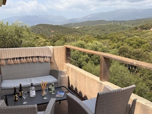 An outdoor seating area with wicker furniture, a table with drinks, and a view of a mountainous landscape.