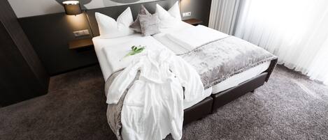 Una cama bien hecha con sábanas blancas y una manta gris, una lámpara de noche y una mesita con lámpara.