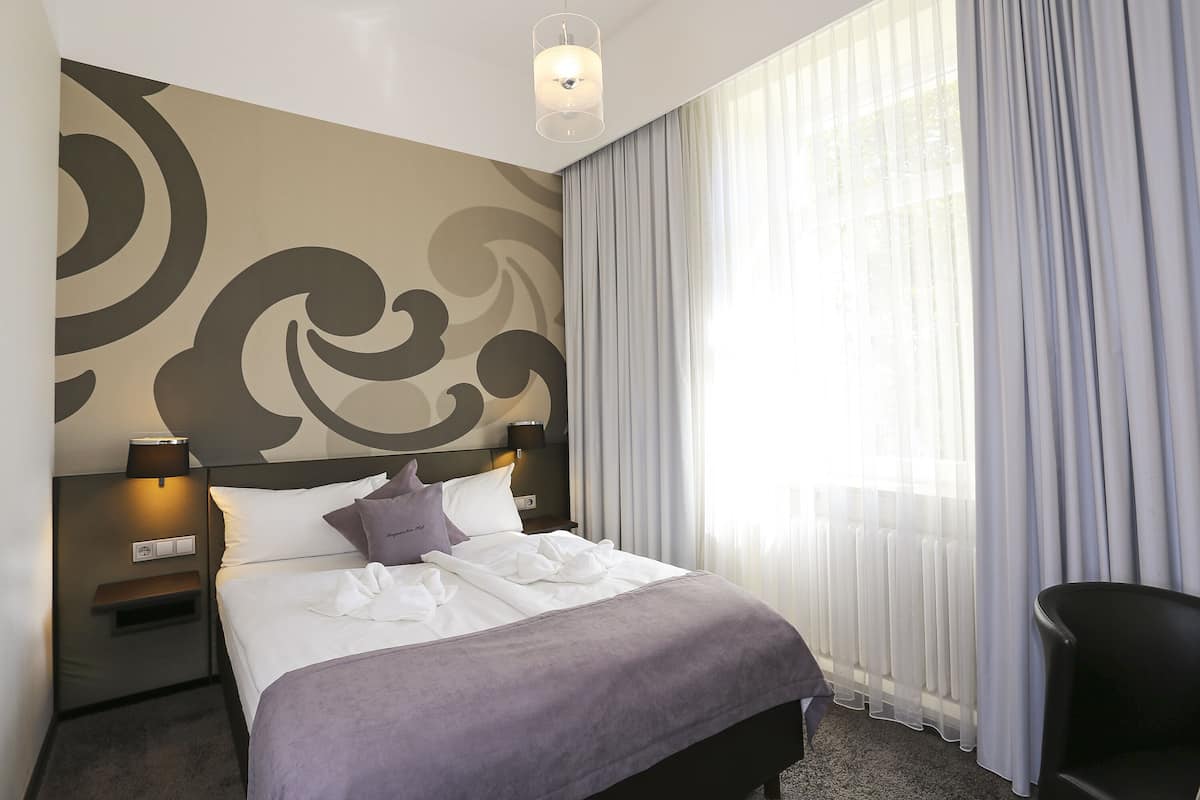 A hotel room with a large bed, a headboard with a decorative wall design, a nightstand with a lamp, and sheer curtains on the window.