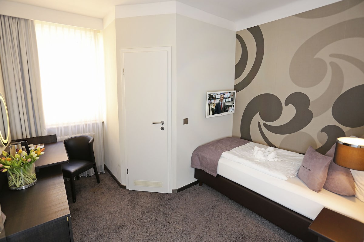 A hotel room with a bed, a desk, a chair, a TV, and a vase of flowers.