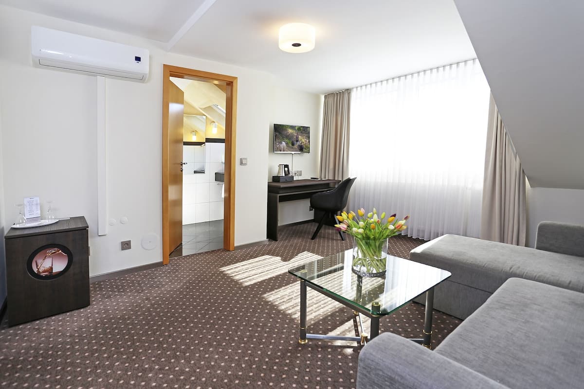 A hotel room with a glass coffee table, grey sofa, and a desk with a chair. There is a small side table with a vase of flowers.