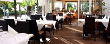 An indoor dining area with tables set for guests, featuring a wooden floor and large windows offering a view of the outdoors.