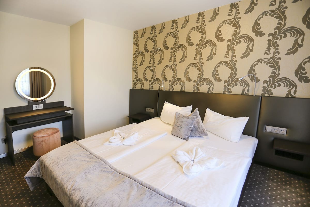 A hotel room with a large bed, a round mirror, a wooden stool, and a patterned wallpaper.