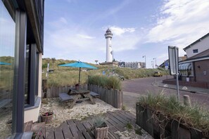 Küstenansicht mit Leuchtturm, einer Holzterrasse, Picknicktisch und Strandstühlen.