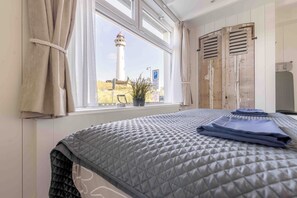 Ein Zimmer mit Bett, einem Vorhangfenster, Blick auf einen Leuchtturm und einer Holztür.