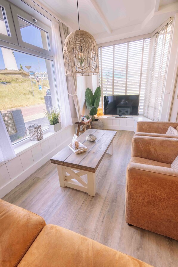 A living room with a wooden coffee table, a sofa, a television, and a large window with blinds. - Top location! Next to lighthouse, beach 50m FLOW (Egmond aan Zee)