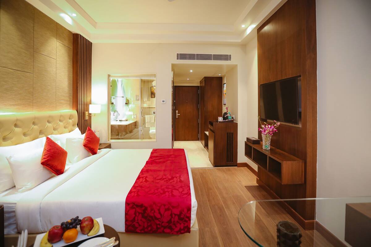 Une chambre d’hôtel avec un grand lit, une télévision, une armoire en bois et une table basse en verre sur laquelle sont disposés des fruits.