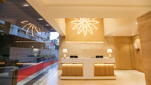 Zona de recepción de un hotel con diseño moderno, que incluye un mostrador de recepción, dos lámparas y una gran lámpara floral en el techo.