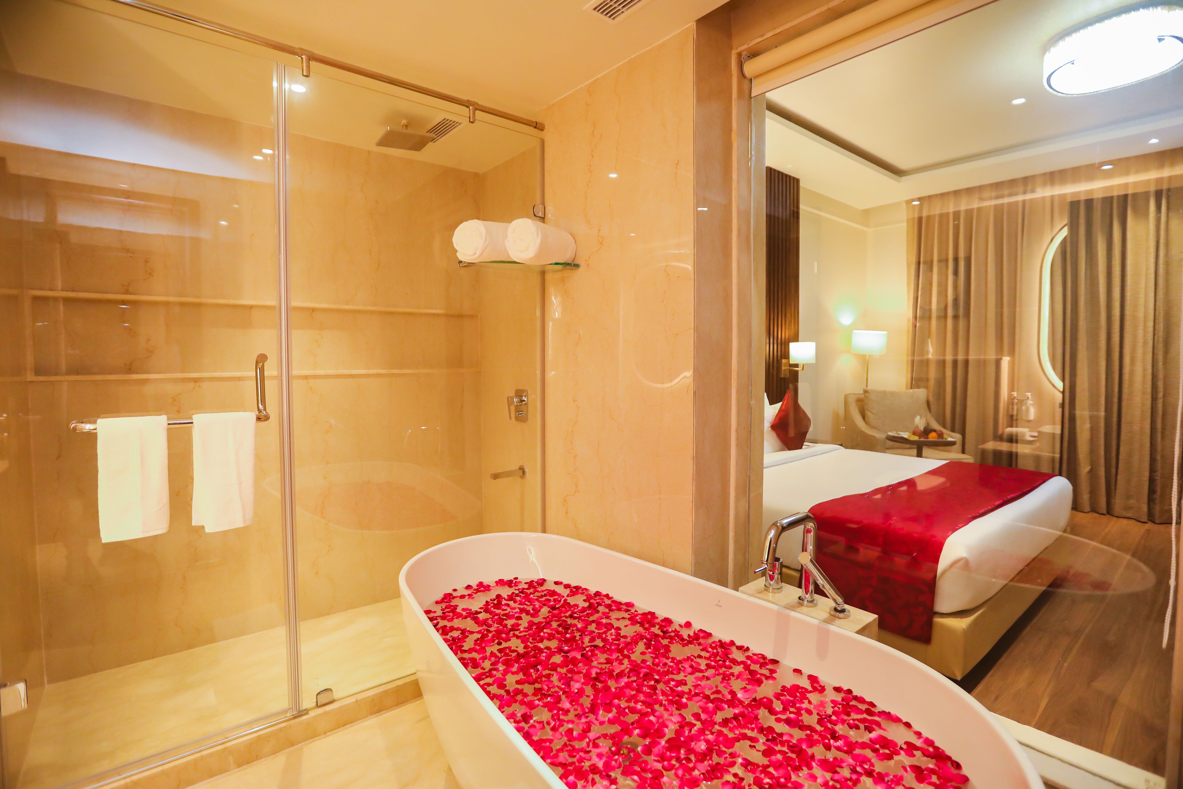 Un baño de hotel con ducha envuelta en vidrio, una bañera llena de pétalos de rosa y una cama con manta roja.
