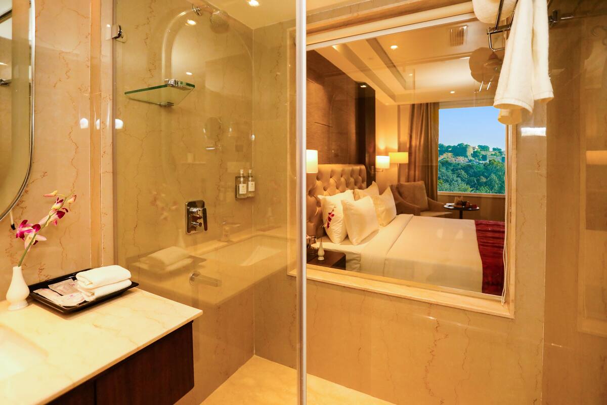 Une salle de bain moderne dotée d’une douche à parois de verre, d’un meuble-lavabo avec des articles de toilette et d’un grand miroir qui reflète une chambre avec un lit et une vue sur la fenêtre.