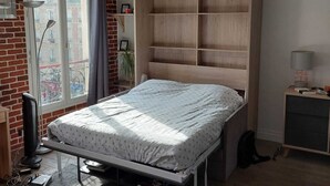 Ett sovrum med en säng, en garderob i trä, ett skrivbord, en lampa och ett fönster med utsikt över byggnader.