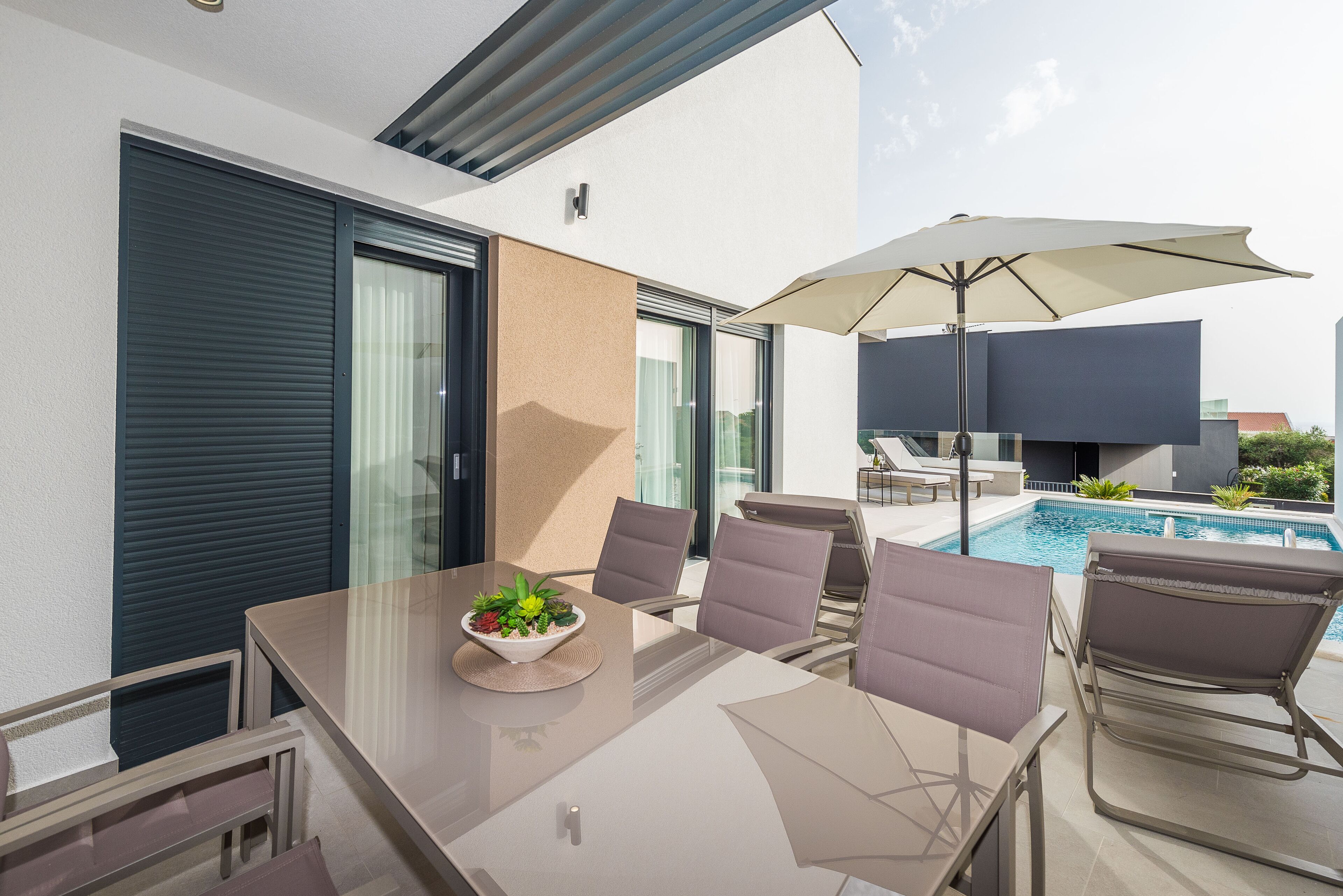 A modern outdoor dining area with a glass table, cushioned chairs, a bowl of fruit, and a pool in the background.