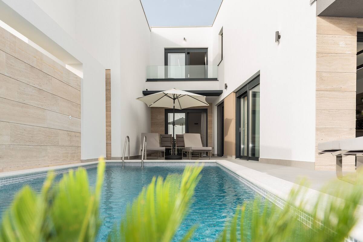 A modern indoor pool area with a clear view of the sky, a seating area with a table and chairs, and a glass balustrade.