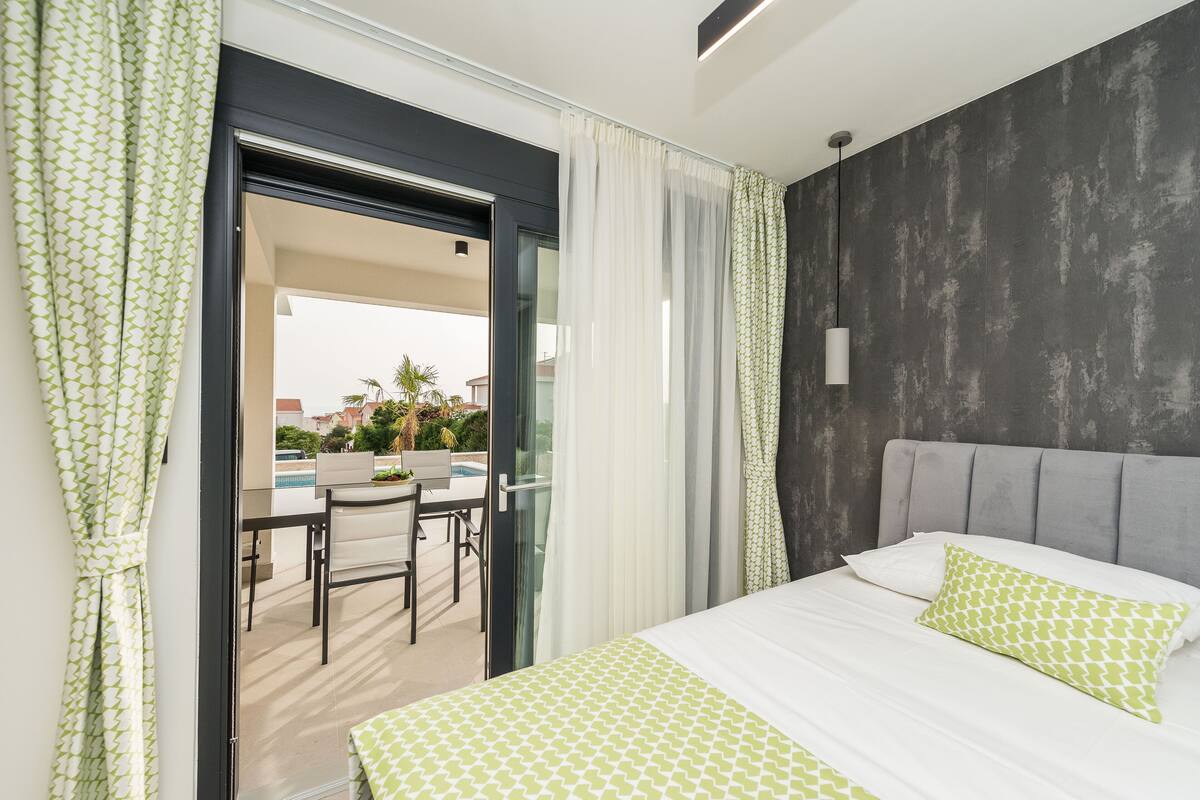 A modern bedroom with a large bed, a grey headboard, and a view of an outdoor pool area.