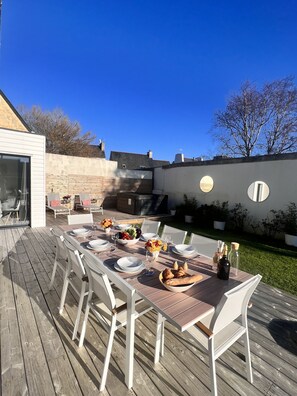 Ein Essbereich im Freien mit einer Holzterrasse, weißen Stühlen und einem für eine Mahlzeit gedeckten Tisch.
