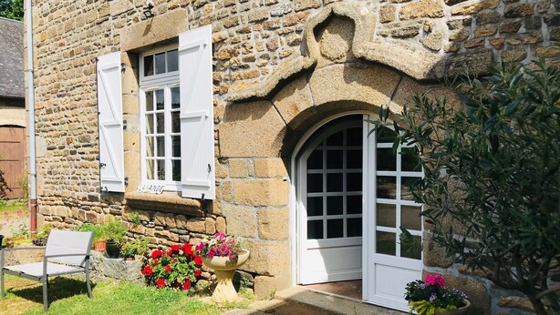 A stone building with two white-framed windows and a door, surrounded by potted plants and a bench.