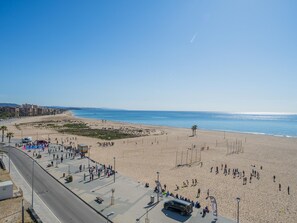 Uma praia com uma estrada paralela, bordeada por palmeiras e alguns edifícios ao fundo.