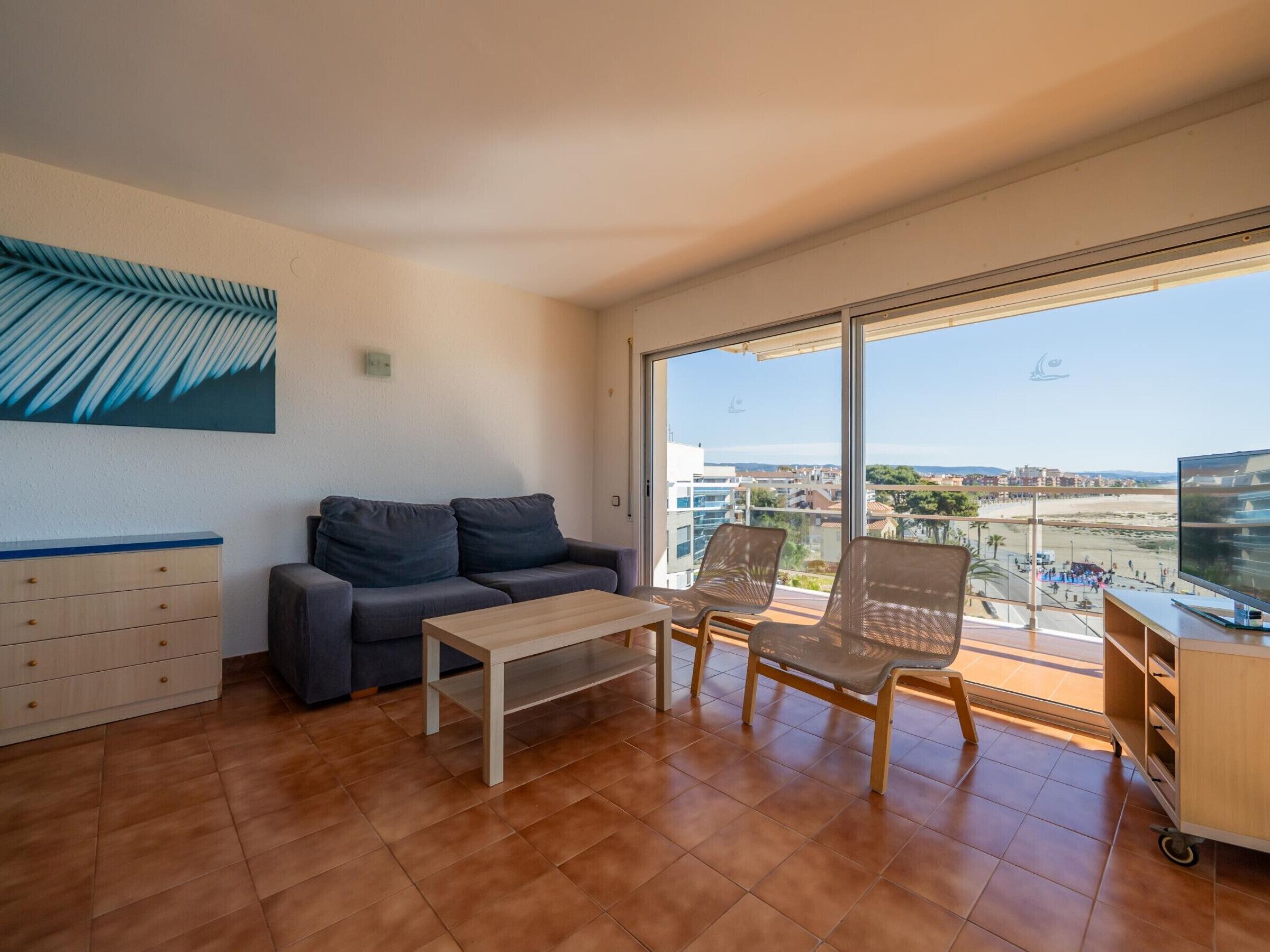 A modern living room with a sofa, a coffee table, and a TV. There is a large window offering a view of the city and beach.