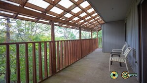 Un balcón techado con barandillas de madera, piso de concreto y dos sillas de metal blanco.