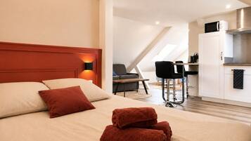 A hotel room with a bed, a desk, a chair, a kitchenette, and a ceiling light.