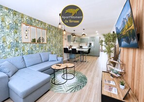 Una sala de estar moderna con un sofá, una mesa de centro y un comedor con sillas. Se observa una cocina al fondo, un televisor montado en la pared y paneles de madera en un lateral.