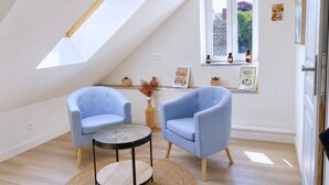 Een kamer met een schuine kap, twee blauwe fauteuils, een klein rond tafeltje, een raam met uitzicht, en een plank met kleine voorwerpen.