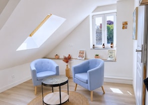 Una habitación con techo inclinado, dos sillones azules, una pequeña mesa redonda, una ventana con vistas y un estante con objetos pequeños.