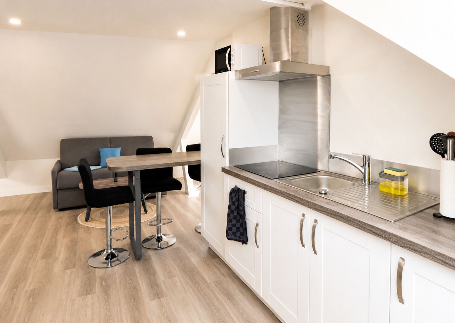 Una cocina moderna con armarios blancos, electrodomésticos de acero inoxidable y un comedor con sofá gris y sillas negras.
