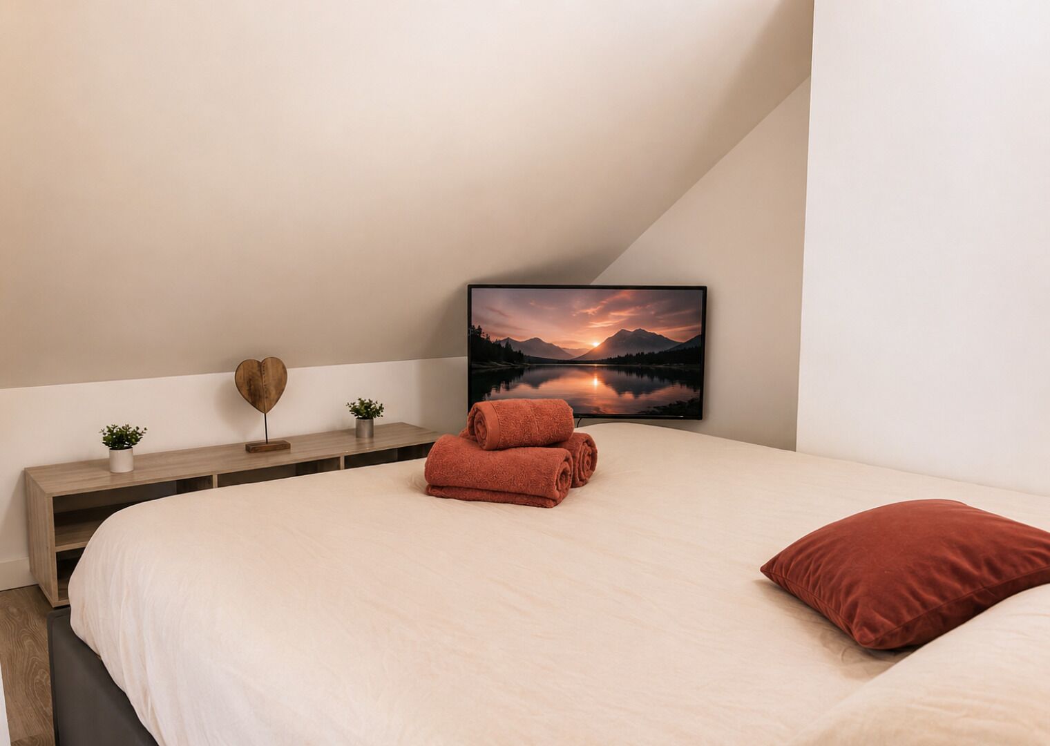 A bedroom with a bed, a TV displaying a mountain landscape, a wooden bedside table with decorative items, and a red pillow.