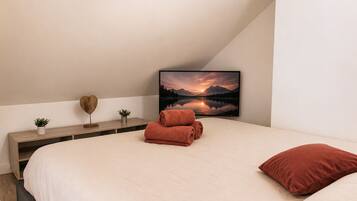 A bedroom with a bed, a TV displaying a mountain landscape, a wooden bedside table with decorative items, and a red pillow.