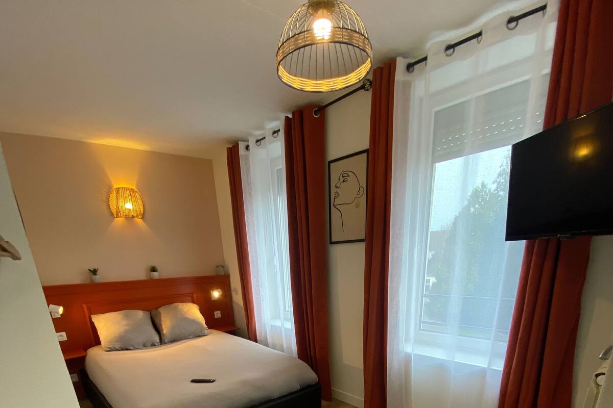 Una habitación de hotel con una cama, televisión, luz empotrada en la pared y ventana con cortinas.