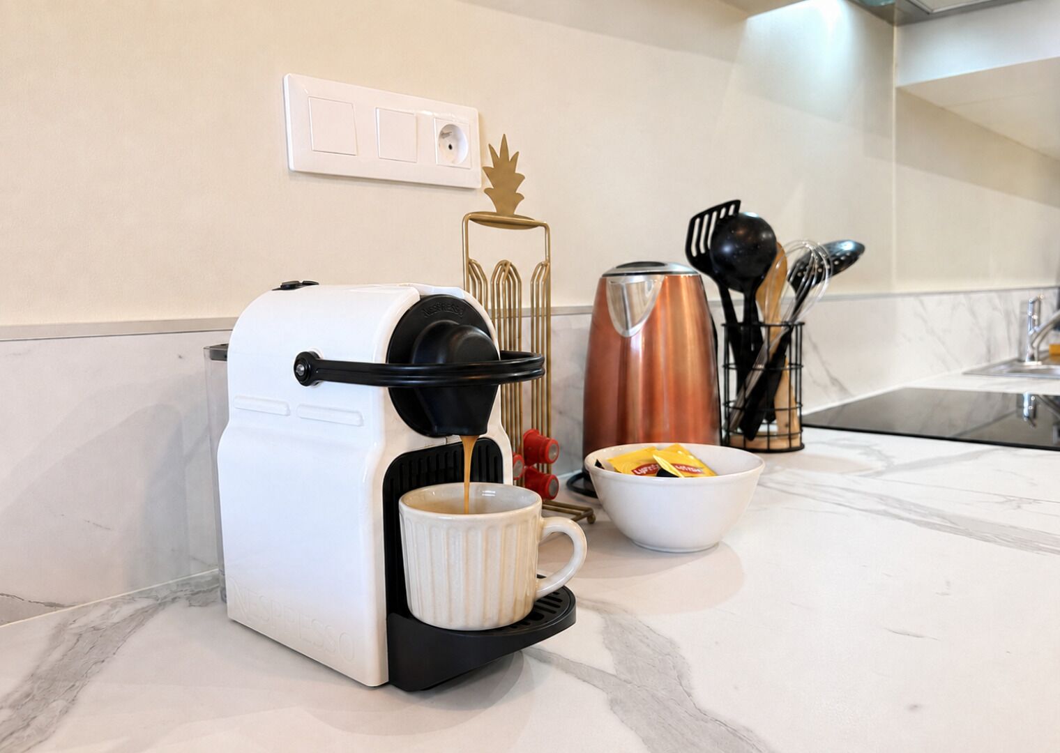 A coffee maker on a kitchen counter with a coffee mug, a bowl of sugar packets, and a coffee pot in the background.