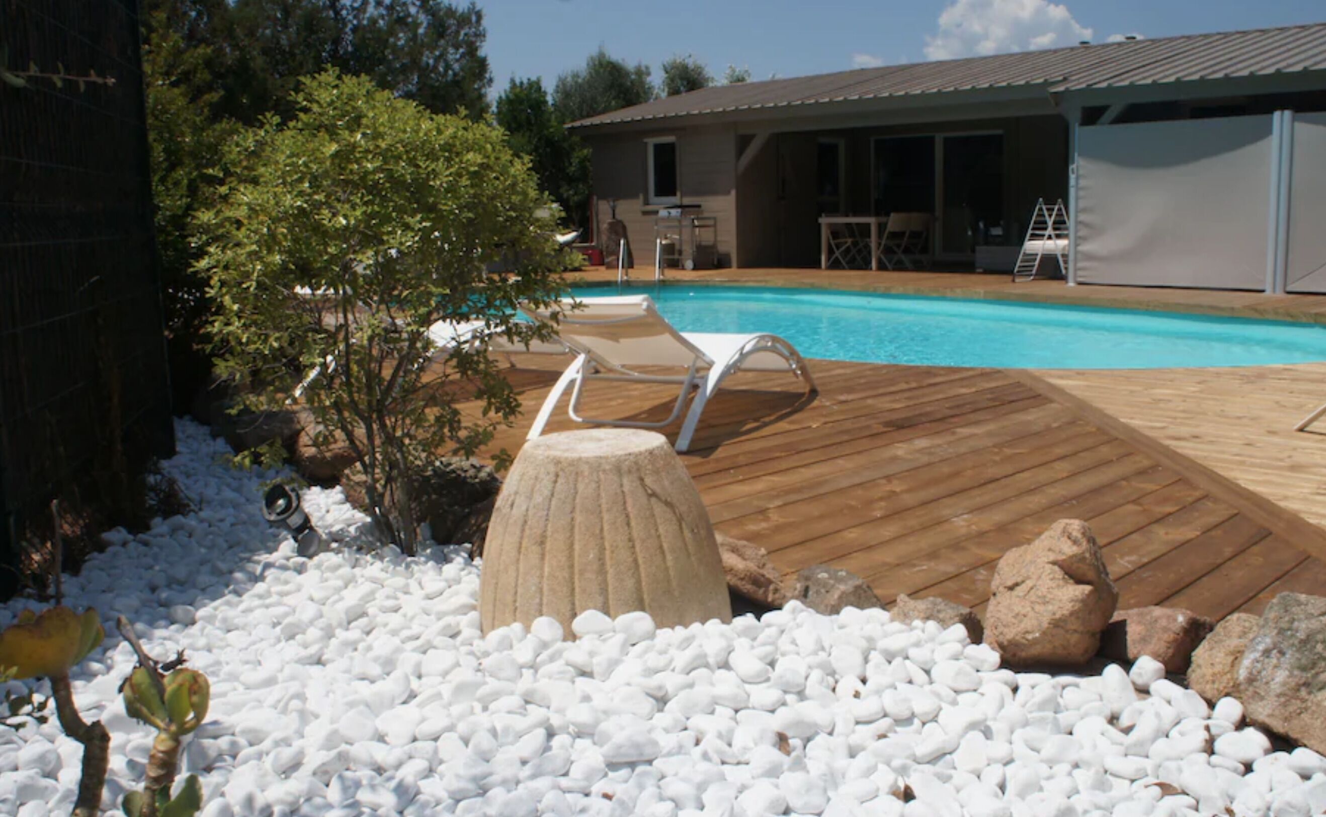 Ein Poolbereich mit Holzterrasse, weißen Kieselsteinen und einem kleinen Baum.