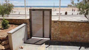 Una entrada cerrada con puerta metálica, muro de piedra y vista a una playa con gente a lo lejos.
