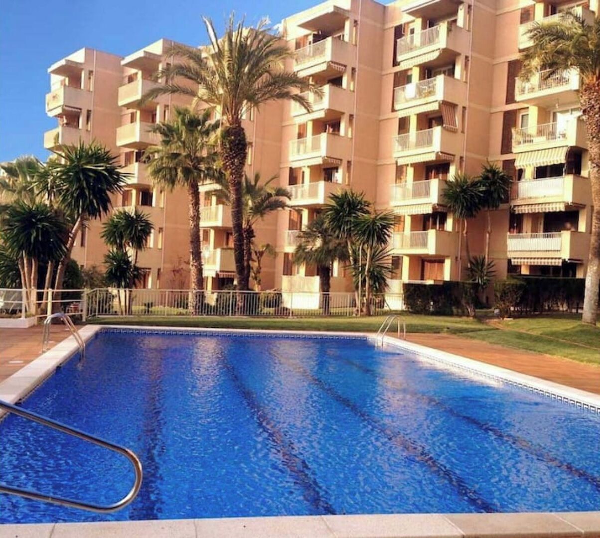 A swimming pool surrounded by a multi-story residential building with balconies, palm trees, and a clear sky.